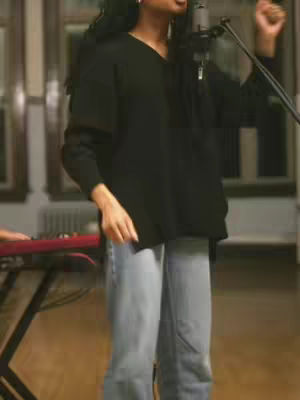A woman with long dark hair wearing a black sweater and light-wash jeans stands in front of a microphone, singing. She is in a dimly lit room with a wooden floor and a keyboard visible in the background. The image is slightly out of focus.
