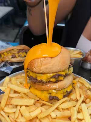 A close-up shot featuring a delicious burger with cheese sauce being poured over it. The burger sits on a bed of fries. Another sandwich is slightly visible in the background.