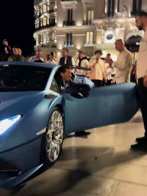 A man in a blue Lamborghini is exiting his car in front of a building at night. A crowd of people stands behind him, and street lights illuminate the scene.
