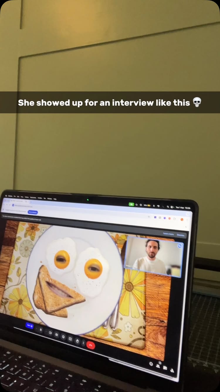 A laptop screen displays a Zoom call with a man in a white shirt, as well as a plate of food in the style of a happy face - with eggs as eyes and toast as a mouth. The background is a wall. The laptop is in front of a green surface