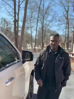 A man in a black jacket stands next to a silver car on a sunny day. The car is on the left, and the man is in the right. Trees with bare branches are in the background.
