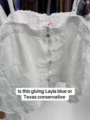 A close-up shot of a white, textured blouse with spaghetti straps. Text overlay at the bottom asks, "Is this giving Layla blue or Texas conservative?" The blouse has buttons down the front and is slightly out of focus, but the fabric looks detailed and slightly crinkled.