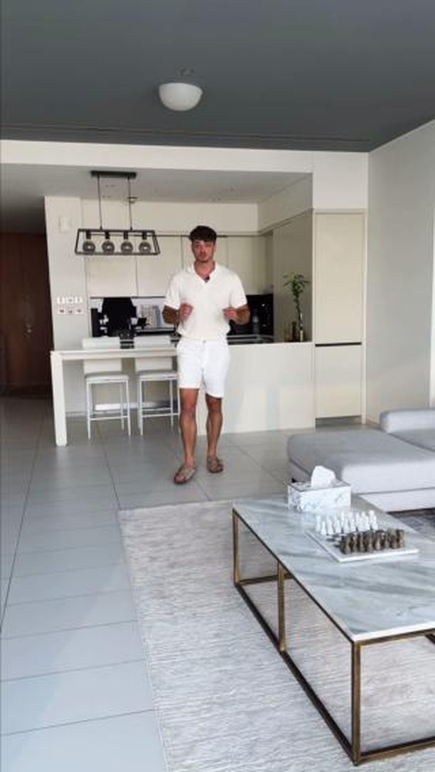 A man in a white polo and shorts walks forward in a modern, well-lit apartment. The background includes a kitchen with white cabinets and bar stools, a dining area, and a gray sofa. A coffee table with a marble top and gold frame sits in the foreground, with a chess set on top. Soft light fills the space.