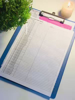 A brightly lit image showing a blue clipboard holding a lined paper, likely for a daily planner or checklist. A pink band is placed behind the clip to hold the pages, and a small green plant sits to the left. There is a softly lit candle in the background to the right.