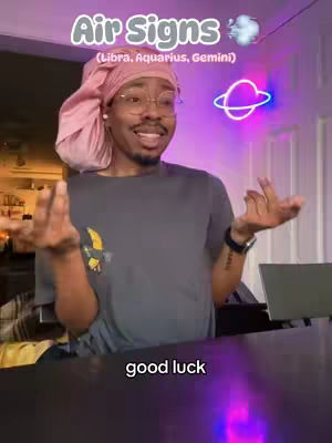 A person with a pink head covering, glasses and a grey t-shirt is centered. Above their head is the text 'Air Signs' in a circular label with a cloud graphic next to it. To the right is a neon sign in the shape of a planet. In the foreground, the text 'good luck' is displayed.