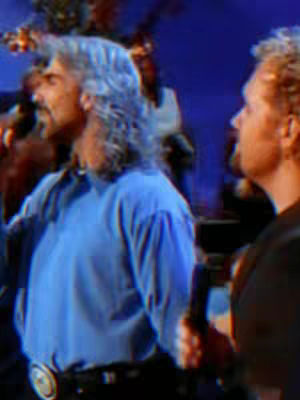 The cover image shows two men, presumably singers, on a stage, captured from the chest up. The man on the left, with long, graying hair and dressed in a blue shirt, is positioned prominently, holding a microphone. To his right, another man with blond hair is partially visible. The background suggests a concert or performance setting, illuminated in blue.