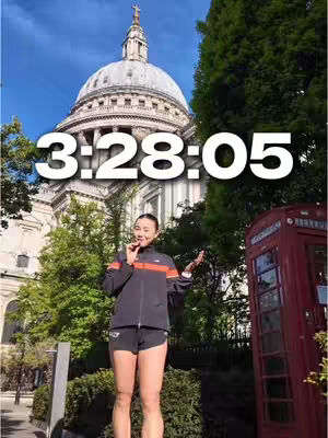 A woman is standing in front of a large building with a domed roof, a clock shows 3:28:05, she is wearing a jacket and shorts, holding something in her hand. Behind her is a building and a red phone booth.