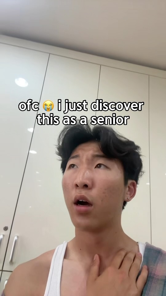A young man with dark hair and an open mouth is the focus, appearing surprised. He wears a white tank top and has a flannel shirt on his shoulder. The background shows white cabinets.