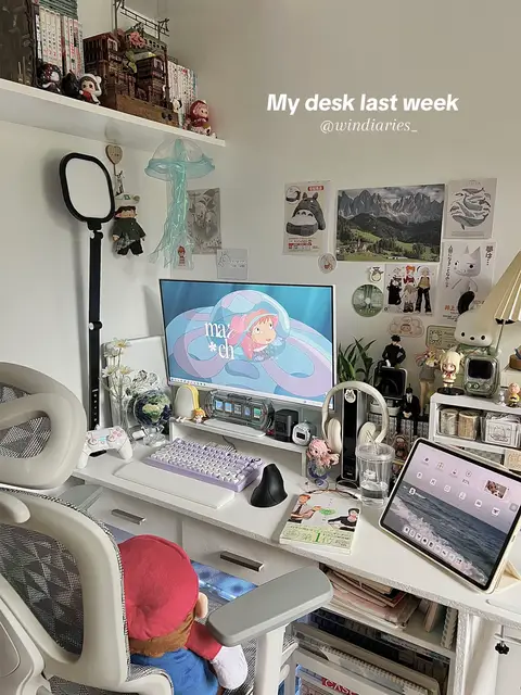 An eye-level shot captures a cozy desk setup, filled with various items. The desk is white and holds a monitor displaying a cartoon image. A keyboard, mouse, and small trinkets are visible. There's a chair with a red Mario doll perched at the edge. The back wall features posters and shelves with more figurines and books, creating a cluttered yet inviting workspace.
