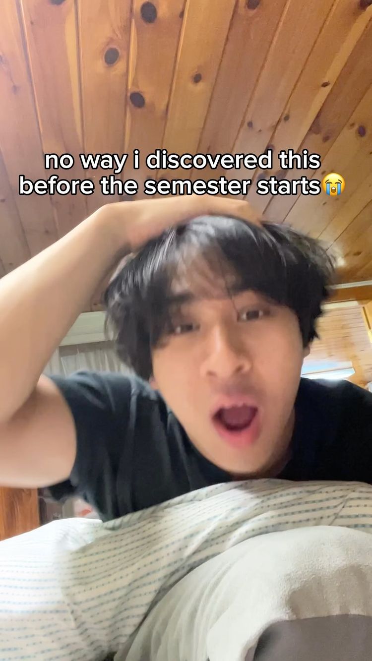 A young man with black hair and an open mouth appears surprised in a selfie-style shot. He's holding his hand to his head, and text overlays the image. The background is a wooden panel.