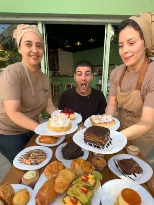 The image shows three people in a well-lit setting. On a wooden table in the foreground, various plates with baked goods are displayed. The two people on either side of the central figure are holding plates with desserts, while the central figure is an open-mouth expression of surprise. The background shows a partial view of a cafe with greenery and outdoor seating.