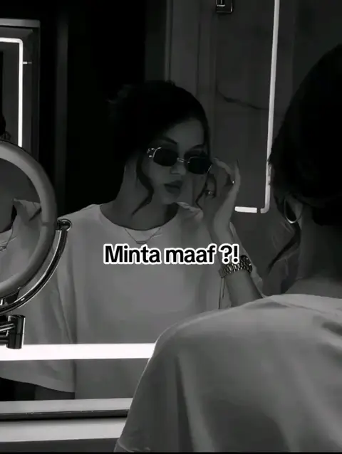 A grayscale image featuring a woman in a bathroom setting. She's reflected in a mirror, wearing sunglasses and a white t-shirt. The text overlay reads "Minta maaf ?!". The overall tone is intimate and modern.
