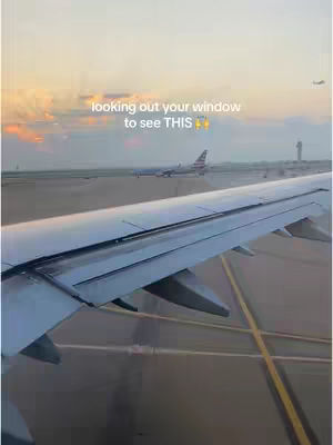 An image from inside a plane, looking out a window. The airplane wing is in the lower right, with the runway and a plane in the background. The sky is a gradient of orange and yellow hues, indicative of a sunrise or sunset.
