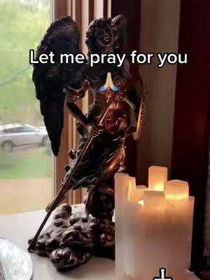 The image shows an ornate statue of an angel with outstretched wings. The angel is holding a staff. In front of the angel, several lit candles are glowing. The text overlay reads, Let me pray for you.' The background shows a window with a car visible from the outside.