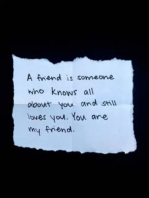 A close-up of a small, torn piece of white paper against a black background. Handwritten on the paper in black ink are the words "A friend is someone who knows all about you and still loves you. You are my friend."