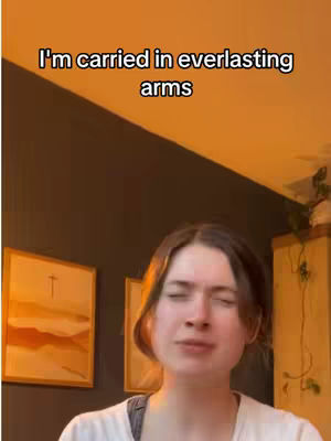 A woman with light skin and dark brown hair is singing in front of a dark wall. The text "I'm carried in everlasting arms" is overlaid at the top. Behind the woman is a painting.