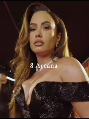 The cover image is a medium shot of the woman, featuring her face and upper body. She has long, wavy brown hair. She is wearing a black, sequined off-the-shoulder dress and a diamond necklace. The background is out of focus, with a warm color scheme, likely from interior lighting. Text overlayed on the image says "8 Arcana".