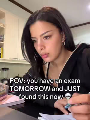 A young woman with long dark hair is looking down with a concerned expression, as if studying. Text overlayed on the image reads 'POV: you have an exam TOMORROW and JUST found this now'. A skull emoji is placed at the end of the text. In the background is a kitchen setting.