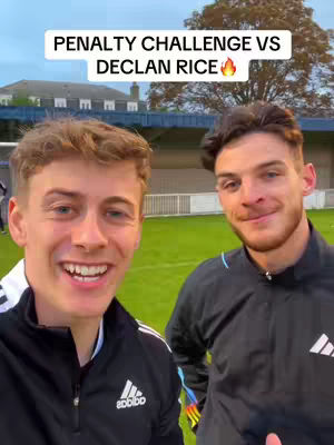 Two men are smiling and posing for a selfie, framed in a medium shot. The man on the left has short, light-brown hair and is wearing a black Adidas jacket. The man on the right has shorter, darker hair and is also wearing a black jacket. The background is out of focus, suggesting a sports field or outdoor setting, with green grass and a building in the background.
