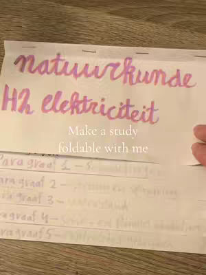 The image shows a close-up of a white paper with handwritten text in a cursive style across it. The top of the paper is folded over. The text is primarily in a purple-pink color. Along the top of the paper, the words 'natuurkunde' and 'H2 elektriciteit' are visible. Below this, the text 'Make a study foldable with me' is handwritten as well. The background is a wooden surface, providing a neutral backdrop to the colorful text.