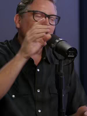 A medium shot of a man speaking into a microphone. He is wearing a black button-down shirt and glasses. His right hand is extended toward the microphone. The background is a solid color.