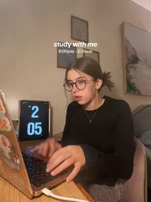 A young woman with glasses sits at a desk working on a laptop, with a tablet displaying the time visible behind her. The scene is likely her room, with decorations on the wall.