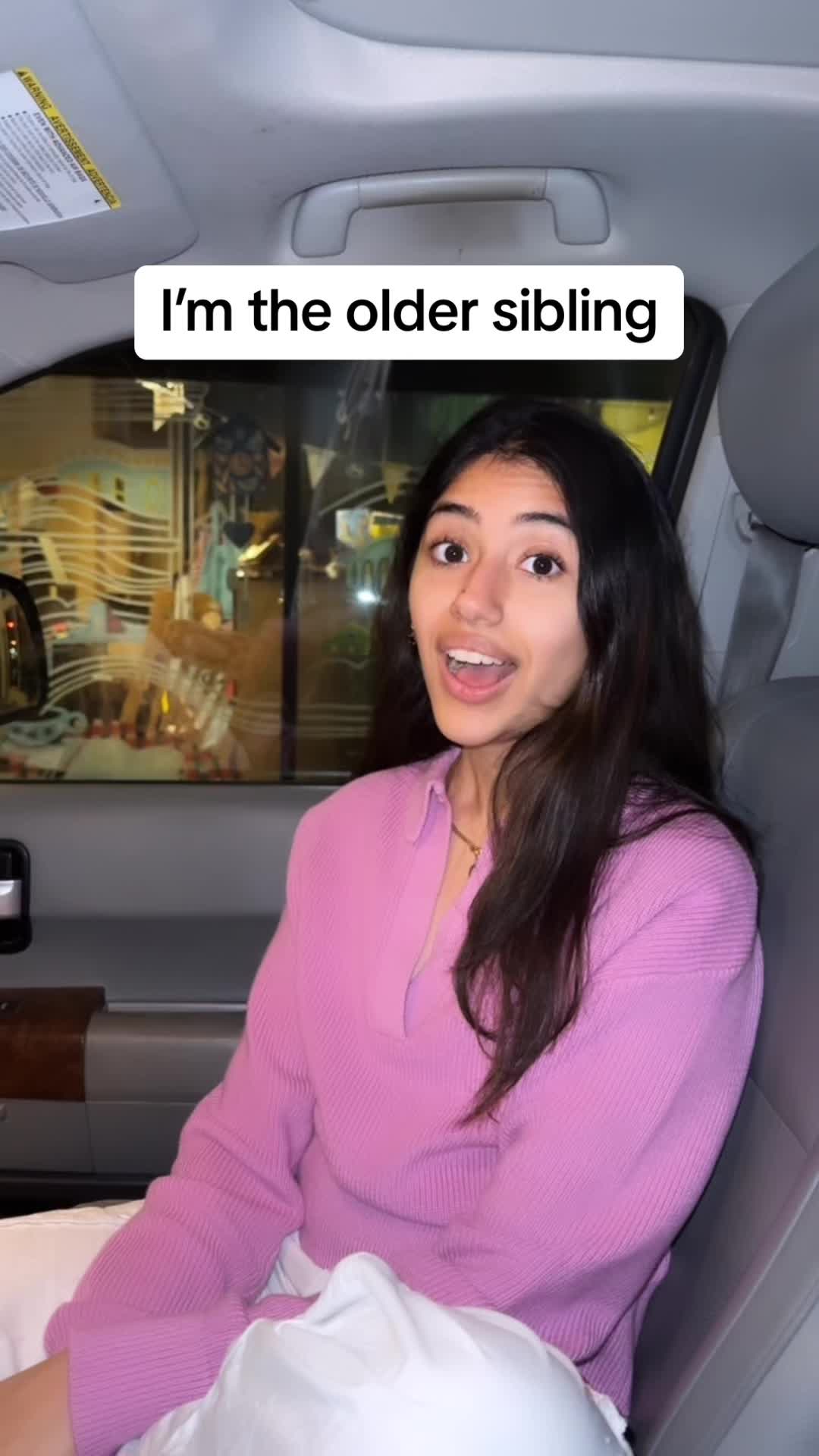 A young woman is seated in a car, likely the driver's seat, with the interior details of the car visible around her. She is wearing a pink top and has long dark hair. Text overlayed on the image reads 'I'm the older sibling'. The woman has an open mouth and seems to be speaking or reacting.