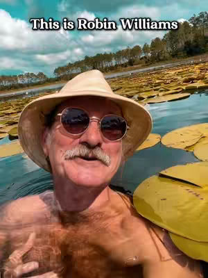 A man in a hat and sunglasses is in a body of water. The water surface is partially obscured by large lily pads. The background has trees and a bright blue sky.