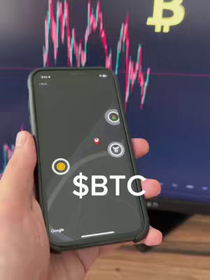 A hand holds a smartphone displaying an app interface, in front of a blurred background showing a stock chart. The phone screen has app icons and the text '$BTC'. The color scheme includes shades of black, white, green and red in the background.