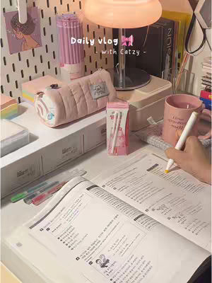 The image shows a well-lit, aesthetically pleasing desk setup, focusing on a study environment. A person’s hand is writing in a notebook, highlighting the study process. Various stationery items such as a pencil case and markers create a cozy and organized workspace. The color palette is soft with shades of pink and white.