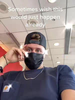 A selfie taken in what appears to be an airport departure area. The man is wearing a black cap, a black face mask, and a Carhartt shirt. The background includes overhead ceiling panels and recessed lighting