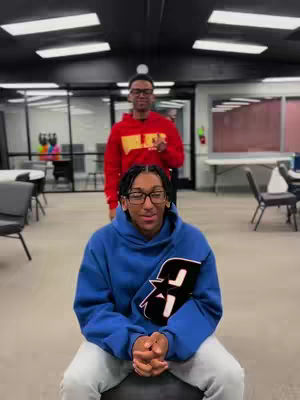 Two young men are in a bright, modern space, likely an office or common area. One is seated, wearing a blue hoodie with a logo, and the other stands behind, in a red hoodie. The seated person faces the viewer while the other guy seems to be looking the same direction. The composition suggests a casual, possibly candid, setting.