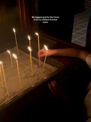 A person placing a lit candle into a sand-filled container. The overall composition is dimly lit, suggesting an indoor, possibly liturgical setting. The color palette is dominated by warm tones from the candles and the surrounding dark environment.