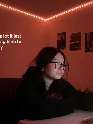 A young woman with glasses is in a room with warm lighting. She is sitting with her arms supporting herself on what appears to be a desk or table. Text is overlaid in the corner of the image, and there are paintings in the background