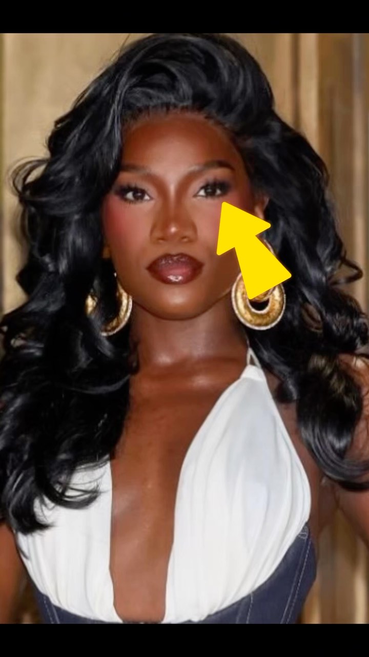 A close-up shot of a woman with dark skin, long black curly hair, and a white halter top. She has full makeup, including dark lipstick, blush, and eyeshadow. Gold hoop earrings are visible, and a yellow arrow is overlaid, pointing to her eye. The background is a blurred beige color.