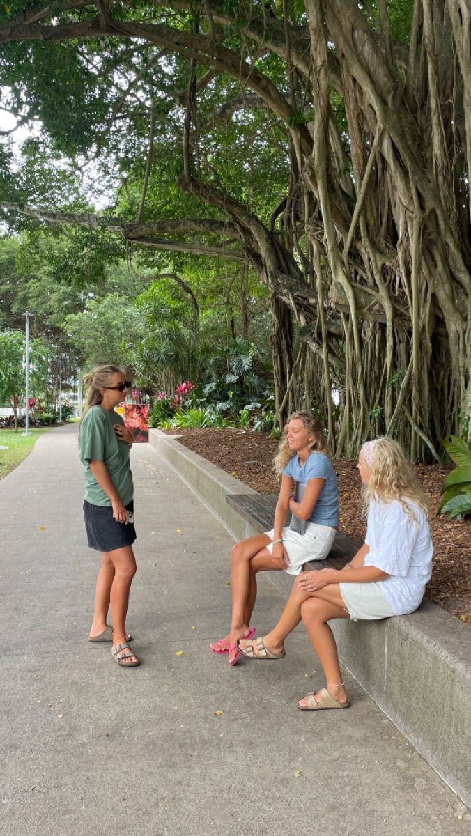 Three young women are conversing under the boughs of a large tree in a park. The woman on the left is standing, wearing sunglasses and holding a brochure, while the other two are seated on a stone bench.  The scene is bathed in natural light, with the green foliage of the tree providing a lush backdrop. The composition is casual and friendly, capturing a moment of connection.