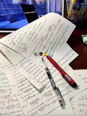 The image shows a detailed, top-down view of a desk covered in handwritten notes filled with mathematical equations, suggesting a study session. Two pens, one red and one black, lie across some of the papers. There are several sheets of paper filled with equations, diagrams, and formulas. In the background, a glimpse of a window and a monitor's screen is visible. There is also a cup containing pens and other supplies. The lighting suggests either a natural or indoor setting, highlighting the papers' content.