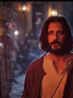 The image shows a man with long, brown hair and a beard, looking slightly off-center. He is wearing a cream-colored shirt under a brown tunic, with a strap visible across his chest. The background is blurred with warm, dim lighting, suggesting an outdoor environment. Overall a medium close-up shot emphasizes the man's facial expression.