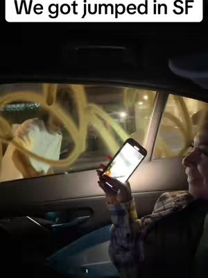 An inside view of a car shows a woman inside filming with her phone. The outside of the car window has yellow graffiti on it. The text overlayed above the car reads 'We got jumped in SF'.