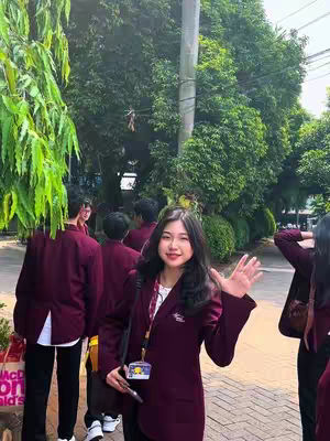 A young woman with long dark hair waves at the camera, standing on a stone path. She wears a dark maroon blazer and a white top, with a small name tag. Behind her, a group of people in similar maroon blazers are visible, blurred, alongside a lush green background. The scene is bright and focused on the woman against a background of greenery and overcast sky.