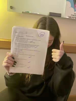 A person is holding up a test paper with a grade of 100 in a thumbs-up pose. The background is blurred and includes a white board, a window, and wooden accents.