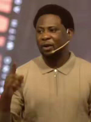 A man with short, dark hair, wearing a beige polo shirt, is speaking with a microphone attached to his ear. He's gesturing with his right hand and appears to be in front of a stage or studio backdrop with soft lighting.
