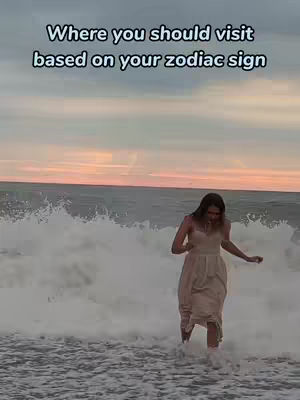A woman in a light-colored dress walks into the ocean with waves crashing around her. The sky has a gradient of colors, from blue to orange and pink, suggesting a sunset.