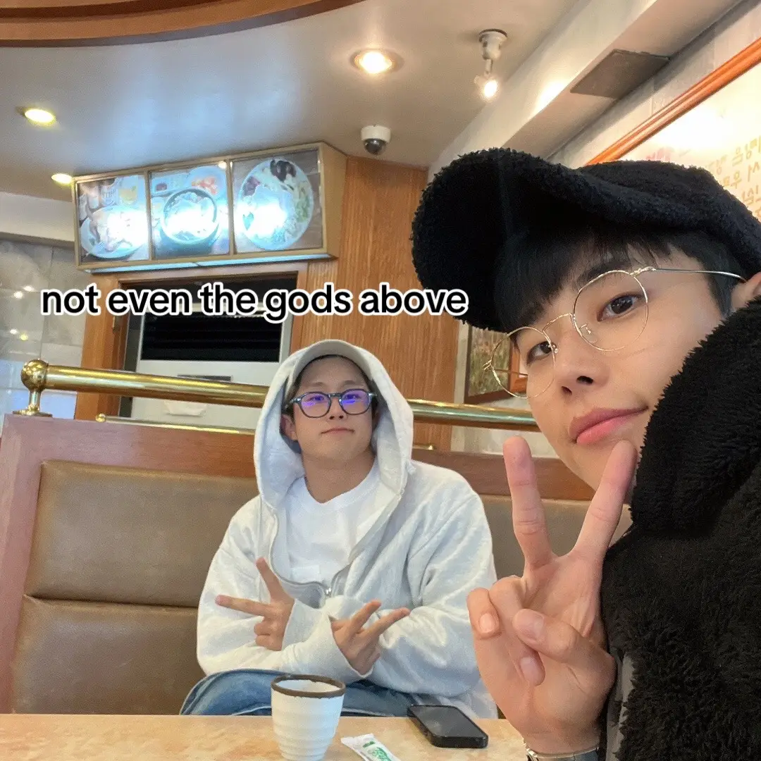 Two young men are featured in a selfie-style shot inside a restaurant. The man on the right is the closest to the camera, smiling and making a peace sign with two fingers up, with his other arm out of frame. He has a black cap, glasses, and a dark furry coat. The man on the left is further back, wearing a white hoodie, glasses, and making the peace sign. The background shows the interior of a restaurant.