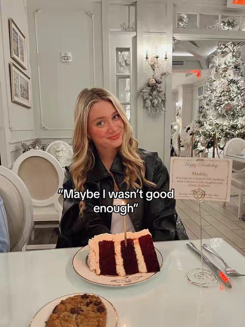 A young woman with blonde hair smiles at the camera while sitting at a white table. In front of her is a plate with a slice of cake, with a lit candle. The setting appears to be a restaurant, with a Christmas tree in the background and decorations that are white themed. There is overlaid text on the image as well.