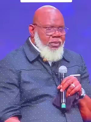 The image shows a bald, elderly Black man with a white beard, wearing glasses and a blue shirt with white polka dots, holding a microphone and looking towards the right. The background is dark blue.