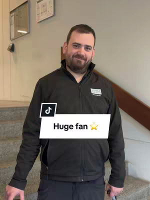 A man in a black jacket stands indoors, possibly in a hallway near stairs. The jacket has a small OTH logo on the front and he is smiling at the camera. A TikTok logo frames a white banner with the words "Huge fan" and a star icon.