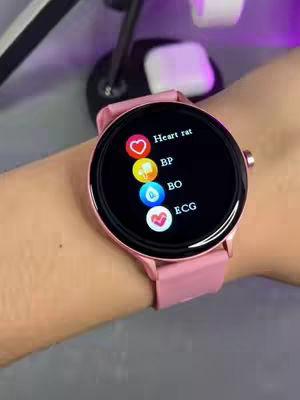 A close-up shot of a pink smartwatch on a person's wrist. The watch face displays various health metrics icons such as heart rate, blood pressure, and ECG, along with their corresponding labels. The background is blurred, but it appears to be an indoor setting with additional elements like other electronic devices.