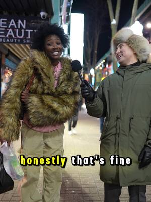 The image captures a street interview setting at dusk. A Black woman wearing a fur coat and khaki pants stands facing a man with a warm hat and olive green jacket, who is holding a microphone. The woman is smiling. A text overlay reads 'honestly that's fine' in a yellow font. Commercial building and people in the blurred background complete the urban scene.