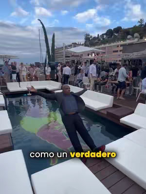 A man in a suit lounges on a pool's edge at an outdoor event. He is in a pool with people enjoying themselves in the background.